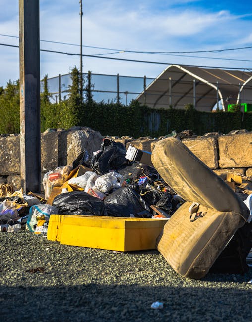 A pile of mixed waste materials is situated on a gravel surface next to a low stone wall. The waste includes black plastic rubbish bags, some torn open revealing paper and plastic waste inside, alongside a yellow plastic bin lying on its side. An old, worn car tire with a dirty beige finish is leaning against the rubble, partially overlapping the yellow bin. In the background, a metal fence topped with a hedge extends across the scene, with utility poles and overhead wires above. A large, semi-circular storage tent or shelter with a metal frame and white covering canopy is visible, along with a blue sky and scattered clouds, suggesting daytime. The scene reflects an area where private waste collection or disposal might occur, associated with a rubbish removal service such as Rubbish Removal Maida Vale, highlighting the accumulation of non-organic waste ready for collection or clearance.