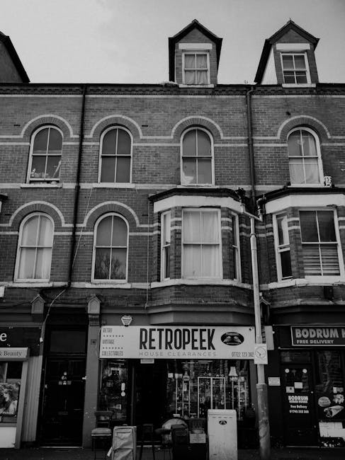 A multi-storey brick building with three levels, featuring large, curved-edged arched windows on the first and second floors, and smaller dormer windows on the top floor with pitched roofs. The building has a traditional red brick exterior with white window frames and decorative detailing around the windows. On the ground floor, there is a retail space with a prominent sign reading 'RETROPEEK' advertising house clearances, accompanied by a graphic of a pair of glasses, indicating it is a secondhand or vintage store. To the right, part of another shop with a sign mentioning 'BODRUM' and free delivery is visible. In front of the building, outdoor seating with a couple of chairs is lined up, and a large, rectangular bin, possibly for waste collection or recycling, is positioned near the entrance. The scene is captured in natural daylight, with some additional detail visible in the reflections on the shop windows. This street-level view is typical for areas where private waste collection or rubbish removal services may operate, supporting the context of independent rubbish clearance services such as those offered by Rubbish Removal Maida Vale.