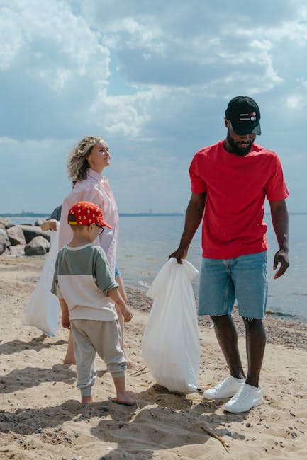 A man and two children participating in a beach clean-up activity during daylight hours. The man is wearing a red t-shirt, ripped denim shorts, white sneakers, and a black cap with a logo, while holding a large white plastic bag. A young boy, wearing a green t-shirt, beige trousers, and a red cap with yellow polka dots, stands nearby holding a second white plastic bag. A woman with wavy blonde hair, dressed in a light-colored or pinkish top, stands slightly behind them, observing the scene. They are on a sandy beach near the water, with a shoreline of large rocks visible in the background. The sky is partly cloudy with patches of blue, creating a natural and casual atmosphere focused on environmental cleanup activities, aligned with private waste collection efforts and rubbish removal services such as those offered by Rubbish Removal Maida Vale.