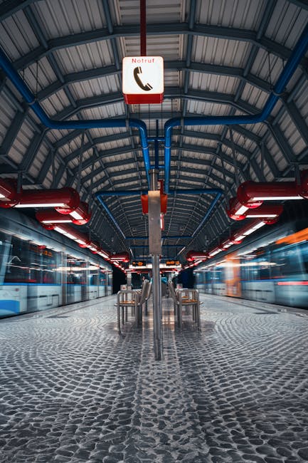 A pile of mixed waste materials situated on a gravel surface in an outdoor area, including black garbage bags, a yellow plastic storage container, a worn beige upholstered armchair with visible fabric texture and some staining, and scattered debris. Behind the waste, there is a low stone wall constructed from irregularly shaped grey stones, with some greenery and trees visible beyond it. In the background, a section of a sports facility or tennis courts with a metal fence and a large white canopy is apparent. The scene is lit by natural daylight, casting shadows on the ground and highlighting the textures of the discarded objects. This image illustrates an instance of excess rubbish accumulation that could be addressed through independent collection or private waste disposal services, such as those offered by Rubbish Removal Maida Vale, to facilitate efficient rubbish removal near Maida Vale station W9.