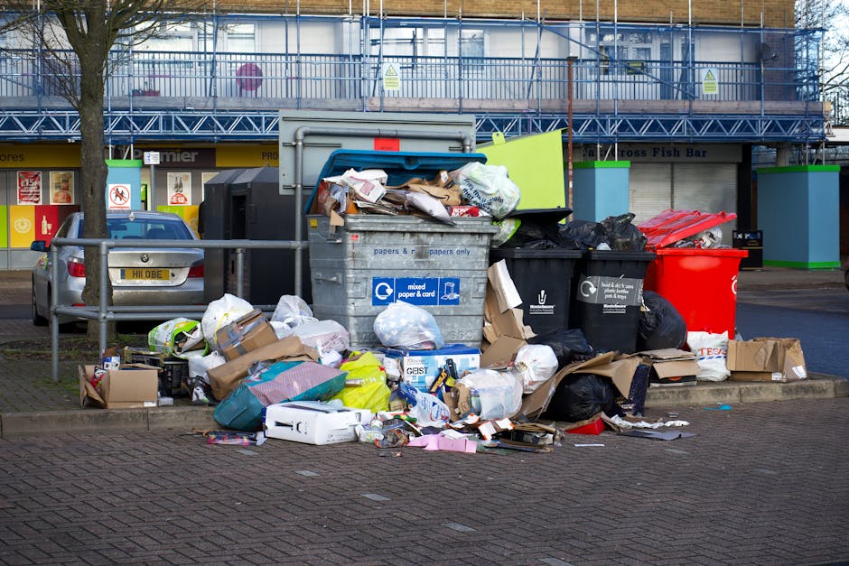 A large collection of mixed household waste and recyclables is piled up next to multiple wheelie bins on a paved area in front of a commercial building. The waste includes cardboard boxes, plastic bags, paper, and packaging materials, some of which are spilling onto the ground. The black, grey, and red wheelie bins are partially open or tipped over, revealing their contents. Behind the waste, there is a blue building structure with safety signs and scaffolding, indicating construction or maintenance work. To the left, a parked silver car is partially visible, with a tree in front of it, and a metal railing separates the waste area from the vehicle and pavement. The scene appears to be an informal or urgent collection of rubbish, highlighting the importance of proper waste management and private disposal services like those offered by Rubbish Removal Maida Vale. The lighting suggests it's daytime with overcast weather, contributing to a neutral, practical atmosphere focused on waste accumulation and organised clearance.