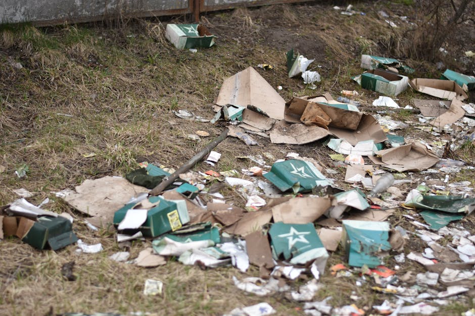 This image shows an outdoor area adjacent to a grassy slope, with a significant accumulation of scattered cardboard boxes, many of which are flattened or torn, revealing their corrugated textures and green, white, and brown color finishes. Some boxes feature printed logos and labels, indicating previous packaging for consumer products. Among the debris are crumpled paper and plastic wrappers, along with a few discarded plastic bottles. A metal pole, possibly a part of outdoor infrastructure or shelving, lies diagonally across the scene, partially covered by the rubbish. The surrounding environment includes a section of a metal fence at the top of the grassy slope, which helps delimit the area. Natural daylight illuminates the scene, highlighting the disorderly arrangement of waste, which appears to be the result of illegal dumping. This scene relates to private rubbish handling and the need for professional waste removal services, such as those provided by Rubbish Removal Maida Vale, to manage and clear such clutter effectively in residential or commercial outdoor spaces.