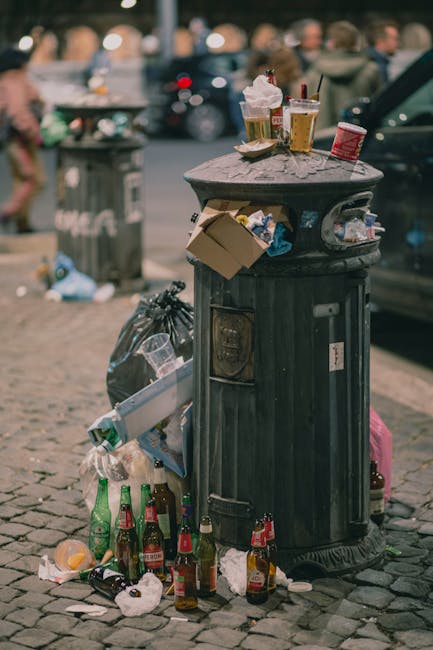 A black, cylindrical public trash bin with a domed lid is situated on a cobblestone street, surrounded by litter including crumpled paper, plastic bottles, and glass beer bottles piled around it and on top of the lid. The bin has a small opening for waste and a slot for newspapers or similar items. On top of the lid, there are various discarded items such as two cups with domed lids containing beverages, one with whipped cream and a cherry on top, and a paper cup, alongside a plastic container. To the left of the bin, a black plastic garbage bag rests against it, containing more waste. The background shows a busy street scene with blurred pedestrians, parked cars, and additional waste bins, suggesting an urban environment during daytime. The scene reflects informal rubbish disposal practices consistent with private waste handling or on-site waste accumulation, aligning with independent rubbish collection services like those provided by Rubbish Removal Maida Vale, which handle debris like this during emergency clearance or scheduled rubbish removal.