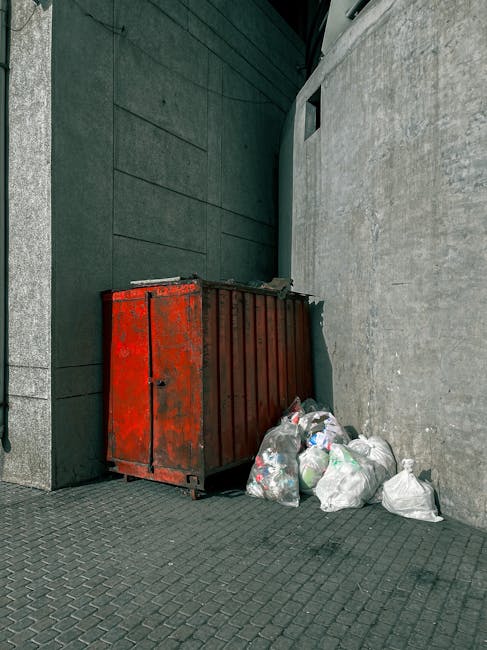 A narrow urban alleyway cluttered with piles of discarded waste and mixed rubbish, including cardboard boxes, plastic bags, and various debris forming a large heap at the rear of the scene. In the foreground, there is a large, weathered wheeled trash bag or container made of dark, durable material, positioned on the gritty, concrete surface that appears stained and uneven. The alley is bordered by aged brick and metal-clad walls, some with graffiti, and features various pipes and utility fixtures attached to the building facades. Bare tree branches extend overhead against a dull sky, contributing to an overall atmosphere of neglect. The scene reflects an environment requiring waste removal services, highlighting the importance of efficient rubbish collection to manage urban clutter and maintain cleanliness, as offered by companies like Rubbish Removal Maida Vale, who provide alternative waste handling solutions apart from local authority collection.