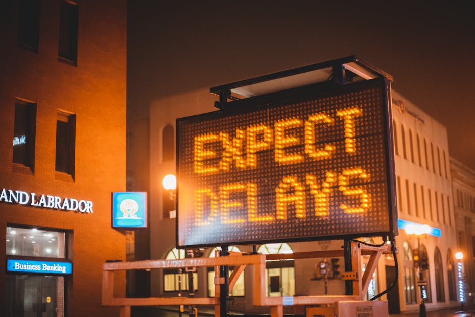 A large electronic road sign displaying the message 'EXPECT DELAYS' in bright orange LED lights, mounted on a metal frame with a black border. The sign is situated outdoors on a city street during nighttime, with surrounding buildings visible in the background. One building on the left has a brick facade with a blue and white sign that reads 'LAND LABRADOR' and another sign below it for 'Business Banking.' The background features a row of streetlights emitting warm glow and several shopfronts with illuminated windows, contributing to a slightly blurred urban scene. The environment suggests an area prone to traffic or service disruptions, possibly related to civic or utility work, and subtly connects to the context of alternative waste handling or service delays for rubbish removal companies like Rubbish Removal Maida Vale.