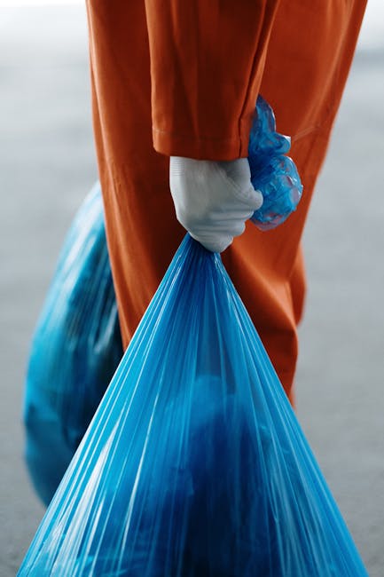 Close-up of a person wearing orange overalls and a white glove, holding a large blue plastic bag filled with waste material. The bag appears to be tightly gathered at the top, secured in the glove's grip, and is situated outdoors on a paved surface. The background is blurred but suggests an open environment, possibly a waste collection point or outdoor processing area. The scene highlights work involved in rubbish collection or disposal, with an emphasis on manual handling of waste in a professional setting. The natural lighting enhances the clarity of the plastic bag's semi-transparent texture and the smooth, sturdy fabric of the overalls. This visual relates to independent or private waste removal services, such as those offered by Rubbish Removal Maida Vale, illustrating the physical aspect of rubbish handling and collection that supports alternative waste management solutions outside local authority services.