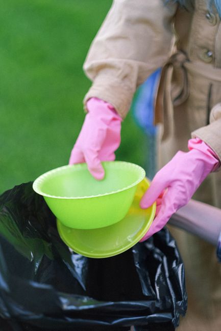 A person wearing a light brown coat and bright pink rubber gloves is holding a small, round, lime green plastic bowl and a matching plate, both with a textured surface. The individual appears to be in the process of emptying or placing these items into a large black rubbish bag, which is positioned on an outdoor surface. The background is blurred but shows a grassy area, indicating an outdoor setting suitable for garden waste disposal or general rubbish collection. The scene suggests an activity related to waste management, possibly involving preparatory steps for rubbish removal or organized rubbish sorting, as handled by companies like Rubbish Removal Maida Vale. Lighting is natural, providing a clear view of the details, including the contrasting colors of waste collection tools and protective gloves against the green and casual environment. The focus on the waste items and the activity aligns with the context of independent waste handling or rubbish collection services in the area, supporting efficient garden waste disposal or general rubbish clearance in accordance with local regulations.