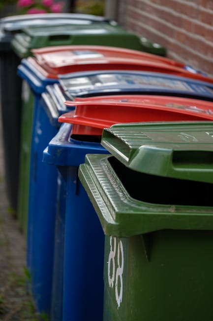 A row of six large wheelie bins positioned on a paved area, primarily made of plastic with textured surfaces. The first bin on the left is dark-colored, marked with a label indicating it is for paper recycling, and features a closed lid. Adjacent to it are five white bins with safely secured lids, all aligned in a straight line, with some labels visible but partially obscured. Next to the bins, there's a black, corrugated metal storage container with a latch, standing on the right side of the frame, and a smaller black stand or sign with white text leaning against the container, indicating disposal guidelines or restrictions. The background consists of dense foliage and overhanging tree branches, creating a natural canopy above the containers. The scene is illuminated by natural daylight, casting soft shadows on the ground. The arrangement and environment suggest a designated area for extraneous waste management or an alternative to council waste collection, managed by Rubbish Removal Maida Vale, supporting private disposal needs within a residential or commercial setting.