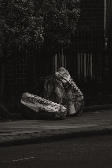 Two large black plastic refuse bags, filled and tied at the top, are positioned on a street pavement near a black metal fence. The bags appear to contain household waste or rubbish, with visible creases and crumpled surfaces. One bag leans slightly against the other, both resting directly on the pavement surface, which has a textured concrete finish. Behind the bags, the fence runs horizontally, partially obscured by dense dark foliage from a nearby hedge or tree, adding a natural backdrop to the scene. The lighting is subdued, creating a shadowed environment, with the overall tone emphasizing an urban setting where private waste collection or rubbish removal services like Rubbish Removal Maida Vale may be used for clearing such waste outside scheduled municipal collections. The image captures the typical scene of roadside rubbish awaiting collection, relevant to discussions on alternative waste handling methods in Maida Vale.