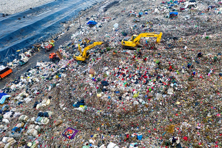 An expansive landfill site filled with layers of mixed municipal waste, including plastic bags, bottles, and household refuse, spread across a large area with uneven terrain. Bright yellow excavators with visible brand markings are actively sorting and moving waste, their large arms extended to shift loads. The waste is intermingled with various colors and textures, from shiny plastics to duller, compacted materials, scattered across the ground and piled in mounds. A partially covered section on the left side is protected by a blue plastic tarp, secured along the edges. The environment appears industrial and utilitarian, with minimal natural features visible, emphasizing the scale and volume of waste processed through private rubbish collection or disposal services like those offered by Rubbish Removal Maida Vale. The scene highlights the physical process of waste management and the importance of effective rubbish removal methods to handle large quantities of refuse in urban settings.