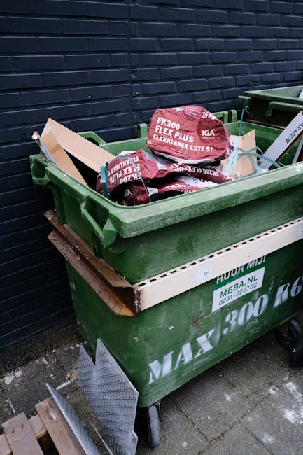 A large green wheeled rubbish bin filled with black and red packaging bags, positioned on a concrete pavement beside a dark brick wall. The bags are branded with red and white labels indicating garden or landscape materials, such as gravel or soil. The bin is partially covered with a white interlocking plastic border, and a wooden plank rests underneath the bin's front edge. In the background, there is a second similar green bin, and the surrounding environment suggests an outdoor storage or waste collection area. The scene relates to private waste disposal services, typical of independent rubbish removal or on-site clearance, with the focus on visible waste packaging and container details relevant to rubbish removal in Maida Vale.