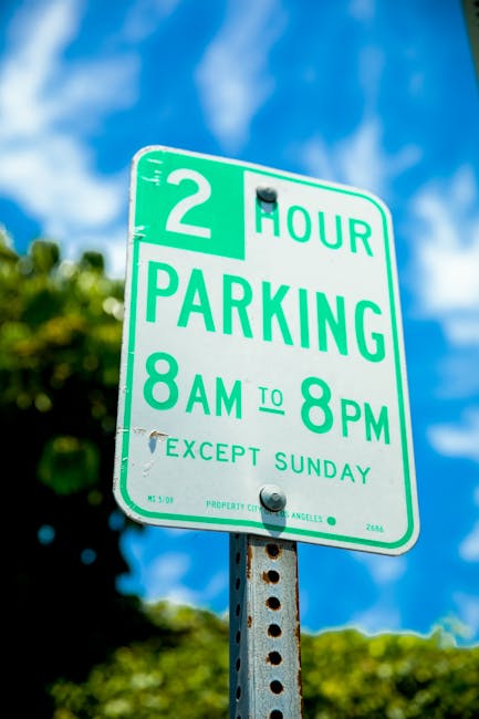 Close-up of a green and white parking regulation sign mounted on a metal pole, indicating a two-hour parking limit from 8 a.m. to 8 p.m., with an exception for Sundays. The sign features bold green text on a white background, with the words '2 HOUR PARKING' prominently displayed in the center, and smaller text below specifying the time restrictions. The background shows a bright blue sky with scattered white clouds and blurred greenery from trees, suggesting an outdoor urban or residential street setting. The sign is positioned in the foreground, with the pole visibly rusted at the bottom, emphasizing its sturdy metal construction. This scene captures standard signage used in local parking management, applicable for private or municipal regulation, relevant to managing vehicle entries during specific hours. The environment indicates a clear, sunny day typical for outdoor parking zones.